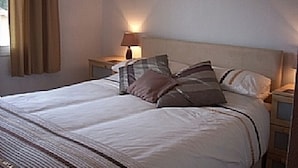 Een net opgemaakt bed met een gestreept deken en meerdere kussens, een bedlampje en een nachtkastje met een lamp.