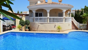 Uitzicht op een zwembad bij een villa in mediterrane stijl, met een gebogen balkon, een gevel van natuursteen en een helderblauw zwembad.