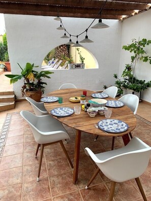 Una mesa de madera redonda con sillas blancas, rodeada de plantas en macetas y un piso de baldosas.