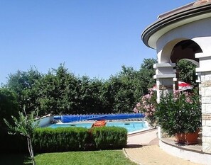 Ein Swimmingpool mit blauer Abdeckung, umgeben von gepflegten Hecken und einem Steinhaus mit Bogen.
