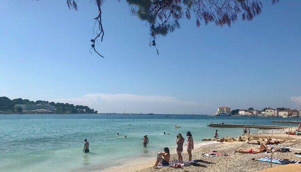 Ein Strand mit klarem Wasser, darin badende Menschen und sonnenbadende Personen, im Hintergrund Gebäude.