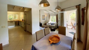 A spacious living area with a dining table, chairs, and a sofa. There is a kitchen visible in the background, and a sliding glass door leading to an outdoor area. - Private villa with mature gardens, 2 minutes walk from sandy beach. (sant lluis)