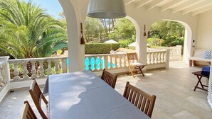 A covered outdoor dining area with a long table and chairs, surrounded by white columns and a white railing. - Private villa with mature gardens, 2 minutes walk from sandy beach. (sant lluis)
