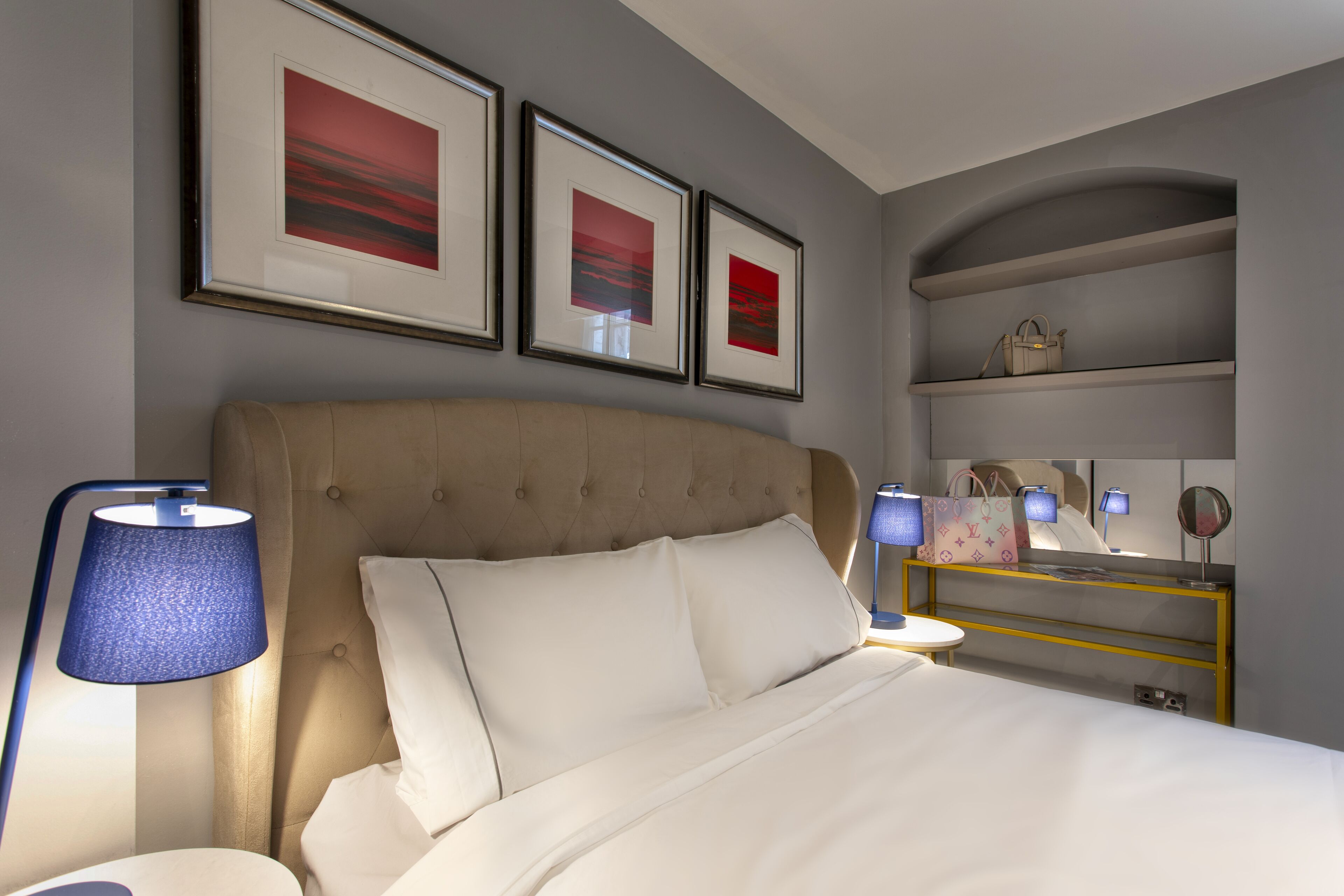 A neatly arranged bedroom with a large bed, a bedside lamp, a vanity with a mirror, and framed artwork on the wall.