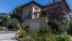 Une maison à deux étages dotée d’un balcon, d’un garage et d’un jardin où poussent différentes plantes et fleurs.