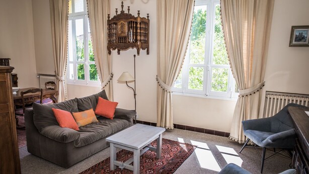 Un salon confortable avec un canapé, un fauteuil et une petite table. De grandes fenêtres sont ornées de rideaux, et on y trouve également un radiateur et une lampe.