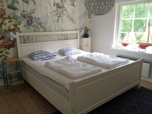 A neatly made bed with white bedding, a wooden nightstand with a lamp, and a floral wallpaper. - For seasonal updates, follow Instagram account sjostuganinsmaland (Solberga)