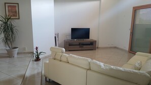 Ein Wohnzimmer mit einem beigen Sofa, einem Fernseher auf einem Holzständer, einer Zimmerpflanze und einer Glastür.