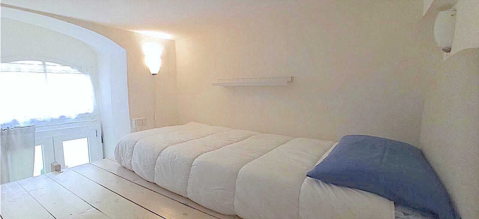 A small, single-bed room with a white bed, a blue pillow, and a wall-mounted light fixture.