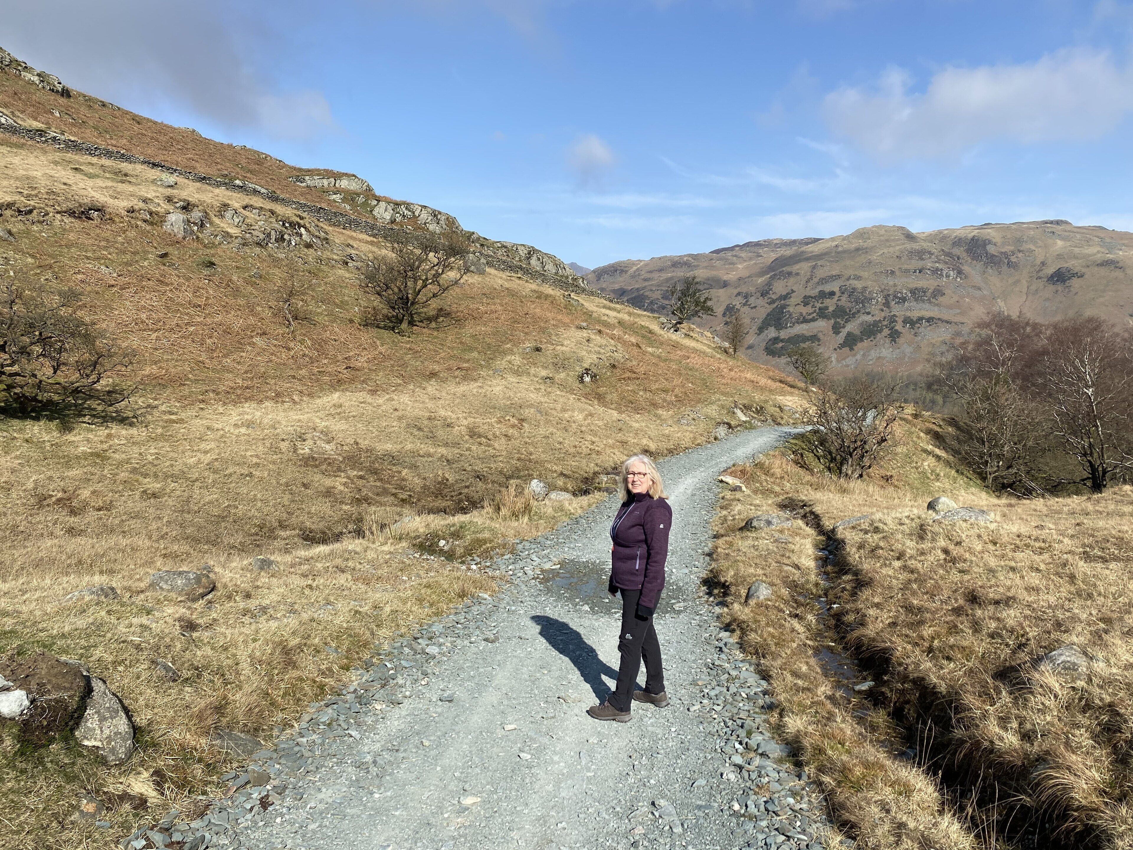 A person standing on a gravel path in a hilly area with sparse vegetation and distant mountains.