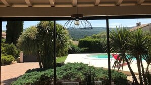 Blick aus einem Zimmer mit großem Fenster auf einen Swimmingpool, umgeben von viel Grün und einer Berglandschaft.