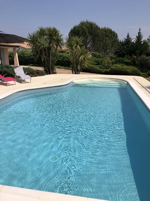 A rectangular swimming pool with clear blue water, surrounded by a tiled deck. There are lounge chairs and a pergola with a roof.