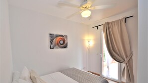 Une chambre à coucher avec un lit, un ventilateur de plafond, une lampe fixée au mur, une fenêtre avec des rideaux et un tableau accroché au mur.
