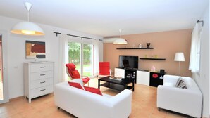Un salon moderne avec un canapé blanc, un fauteuil rouge et une table basse noire. On y trouve un téléviseur posé sur un meuble noir et une armoire blanche.