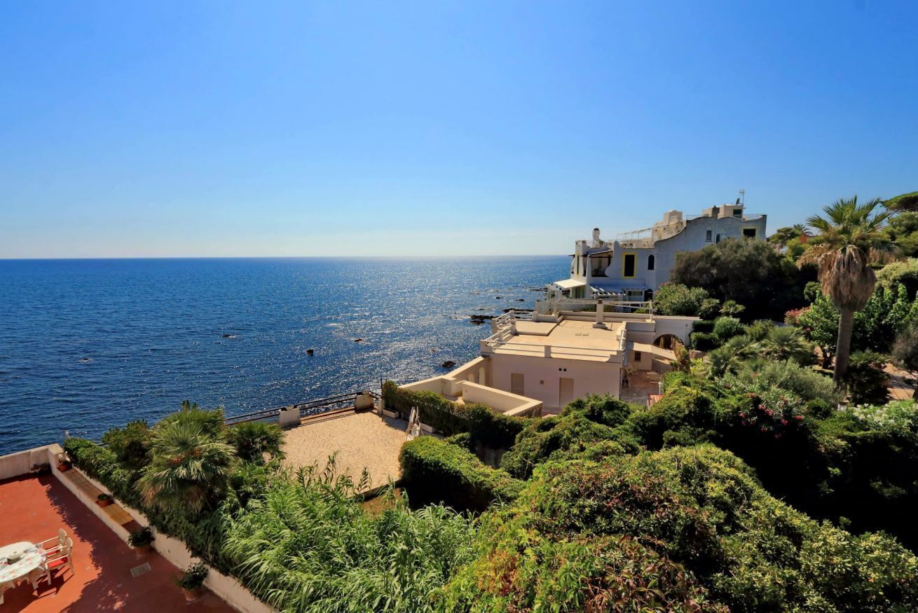 A coastal property with a view of the sea, a patio with outdoor furniture, and lush greenery.