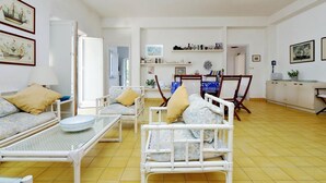 A room with a bed, a sofa, a dining table, and a chair. There are framed pictures on the walls and a shelf with bottles.