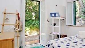 A room with a bed, a wooden desk, a shelf with books and a framed picture, and a door leading to a garden.