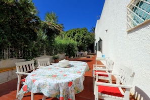 Une terrasse avec une table ronde et des chaises blanches, entourée de verdure et bordée d’un bâtiment blanc.