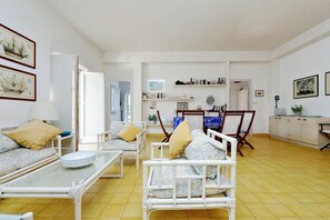 A room with a bed, a sofa, a dining table, and a chair. There are framed pictures on the walls and a shelf with bottles.