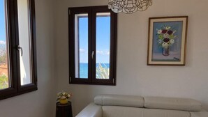 Une pièce avec un canapé en cuir beige, une petite table sur laquelle est posé un vase de fleurs, un tableau encadré représentant des fleurs et une fenêtre donnant sur la mer.