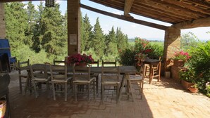 Un patio al aire libre con una mesa y sillas de madera, rodeado de plantas en macetas y vistas al campo.