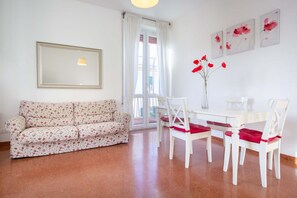 A living room with a floral sofa, a dining table with chairs, and a vase with red flowers.