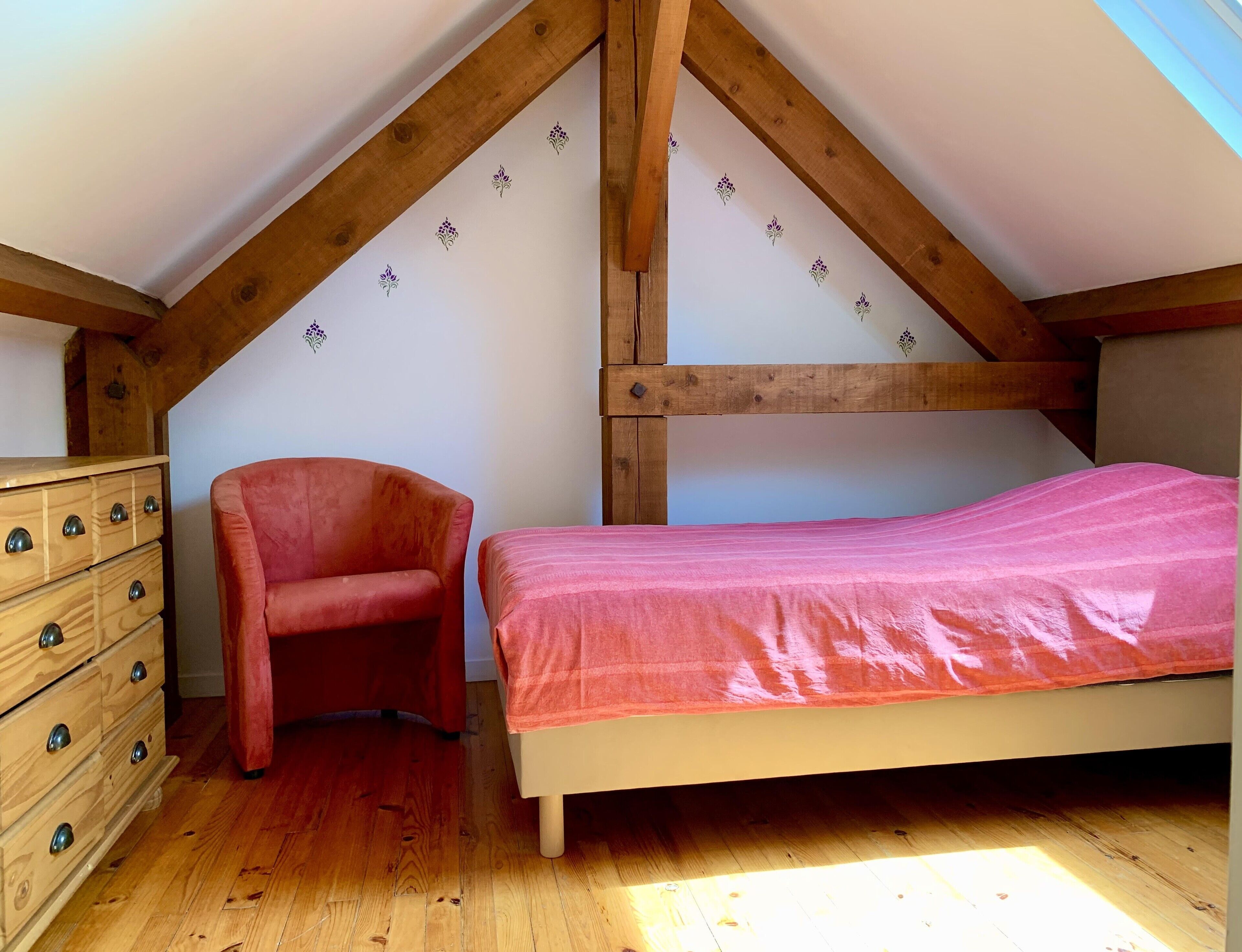 Une chambre avec un plafond en bois, un lit recouvert d’une couverture rose, une chaise rouge et une commode en bois dotée de tiroirs.
