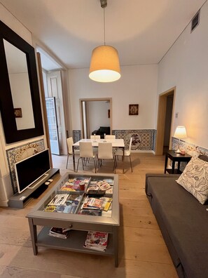 Una sala de estar con televisor, mesa de centro con revistas, sofá y un comedor con sillas y una mesa.