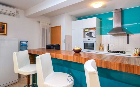 Uma cozinha moderna com bancada de madeira, banquetas brancas, forno embutido e painel de fundo verde-água.