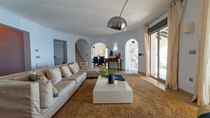 A modern living room with a beige sofa, a white coffee table, and a large brown rug. There is a staircase leading to an upper floor, and a dining area is visible in the background. - Villa Rosh, A luxury, 3 floor villa with majestic sea views this large villa is perfect for a large ... (Xàbia)