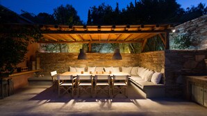 Un patio exterior con pared de piedra, pérgola de madera y zona de estar con cojines.