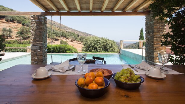 Una mesa de madera con cuencos de fruta y copas de vino, con vistas a una piscina y un paisaje montañoso.