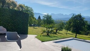 Une terrasse extérieure dotée de chaises modernes, d’une piscine et offrant une vue sur un paysage verdoyant, avec des arbres et des montagnes à l’horizon.