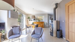 Un salon moderne avec deux fauteuils gris, une table basse en bois et un téléviseur placé à gauche. On aperçoit une cuisine avec une grande fenêtre et une cuisinière au fond.