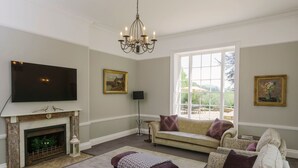 A living room with a fireplace, a television, a chandelier, and a large window. - Magnificant 9 Bedroom Georgian House, indoor pool, cinema room, Sleeps 18-22 (Barnstaple)