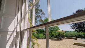 A view through a window of a gravel patio with a table and chairs, surrounded by greenery and a body of water. - Magnificant 9 Bedroom Georgian House, indoor pool, cinema room, Sleeps 18-22 (Barnstaple)