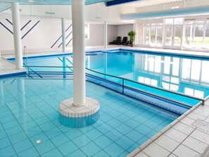 Una piscina interior con agua azul cristalina, suelo de baldosas y una columna blanca en el centro.