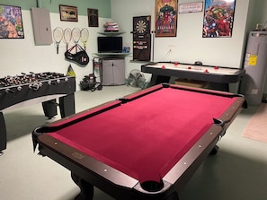 A pool table with a red felt surface, a foosball table, and a dartboard in a room with green walls and various sports memorabilia.