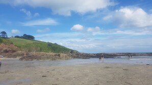 A beach with a grassy hill in the background, rocky shoreline, and people walking along the shore. - Beautiful Country Cottage (Looe)
