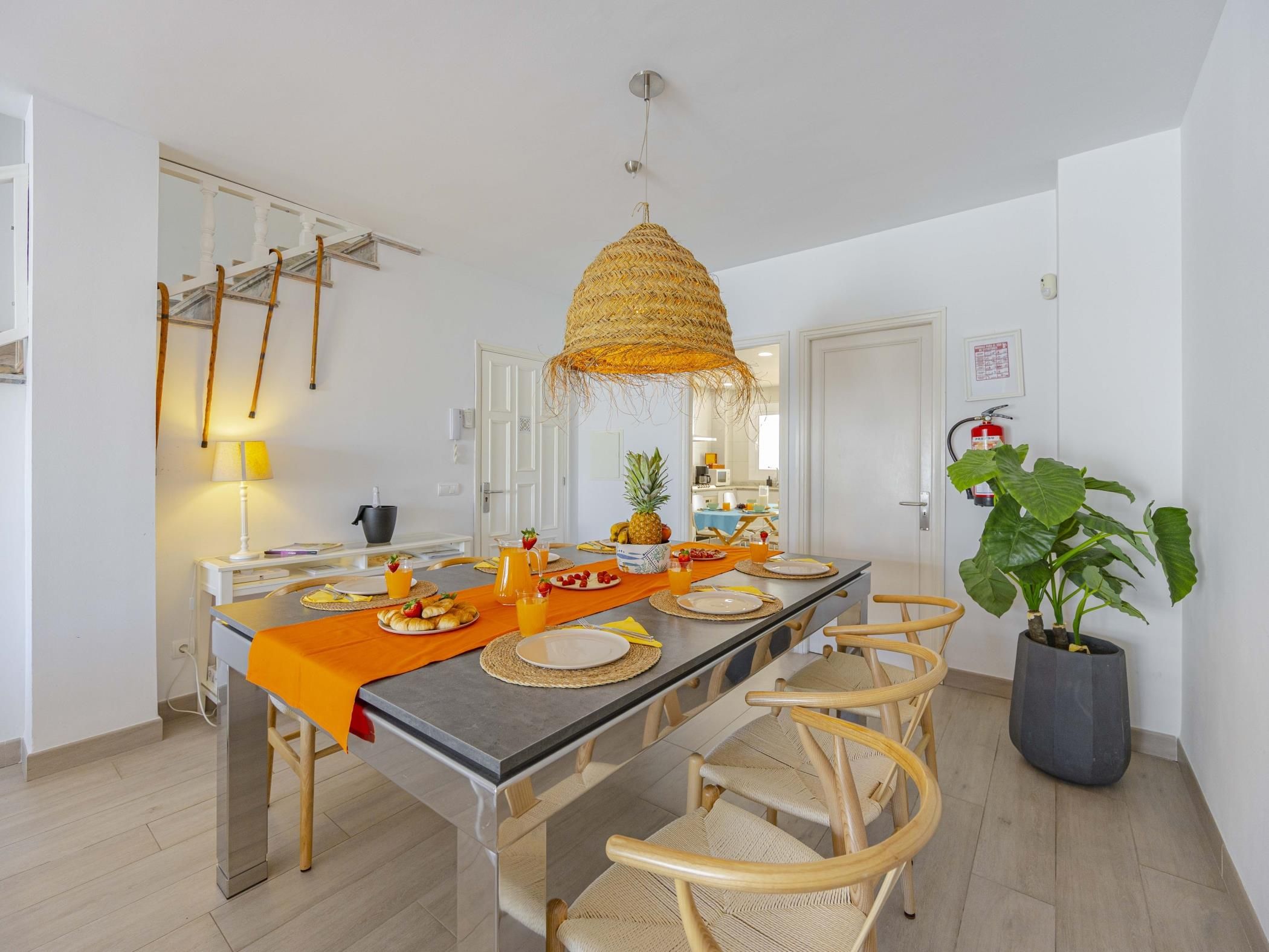 A dining area with a long table, chairs, a woven hanging light fixture, and a potted plant.