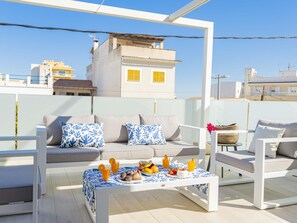 Eine Dachterrasse mit einer Sofagarnitur, einem Tisch mit Obst und Getränken und Blick auf Gebäude im Hintergrund.