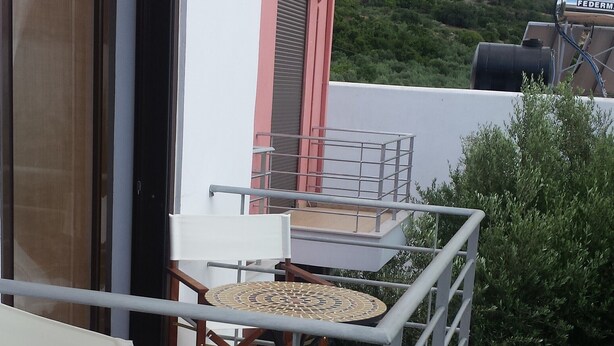 A balcony with a table and chairs, a window with blinds, and a view of a hilly landscape.