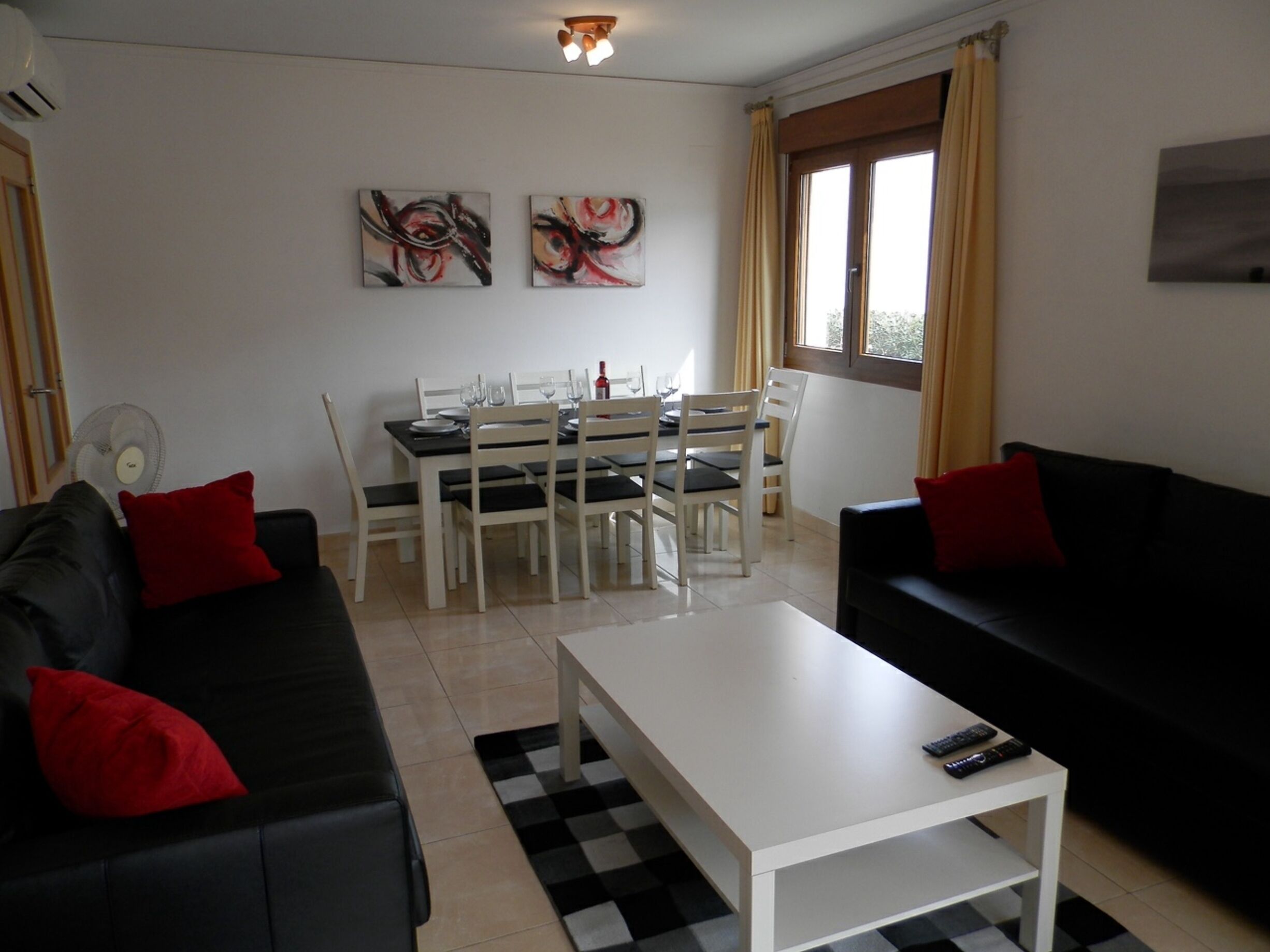 Ein Wohnzimmer mit einer schwarzen Couch, einem weißen Couchtisch und einer schwarzen Couch mit roten Kissen. Es gibt einen Esstisch mit Stühlen und ein Fenster mit Vorhängen.