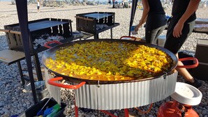 Ein großes Kocharrangement im Freien mit einer Metallpfanne voller Speisen, umgeben von Tischen und Stühlen.
