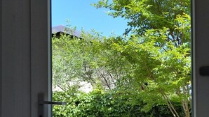 Eine Tür, die in einen Innenhof führt, mit einer Steinsitzbank, einem Blumentopf und Blick auf Grünflächen sowie ein Gebäude im Hintergrund.