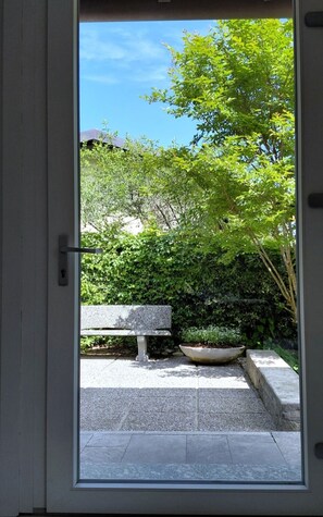 A door leading to a courtyard with a stone bench, a planter, and a view of greenery and a building in the background.