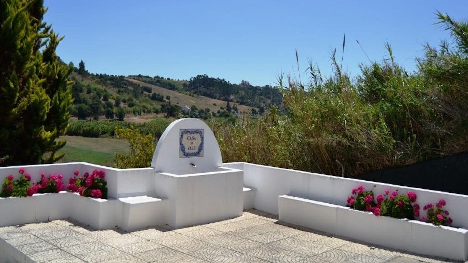 A white outdoor terrace with a sign reading "CASA DE VALE" surrounded by pink flowers and a view of a green valley.