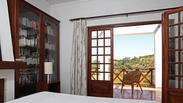 A bedroom with a large bed, a wooden bookshelf, a chair, and a balcony with a view of the countryside. - FIFTH WITH PRIVATE POOL AND COURT TENNIS A 30 LISBON M (Vale do Paraíso)
