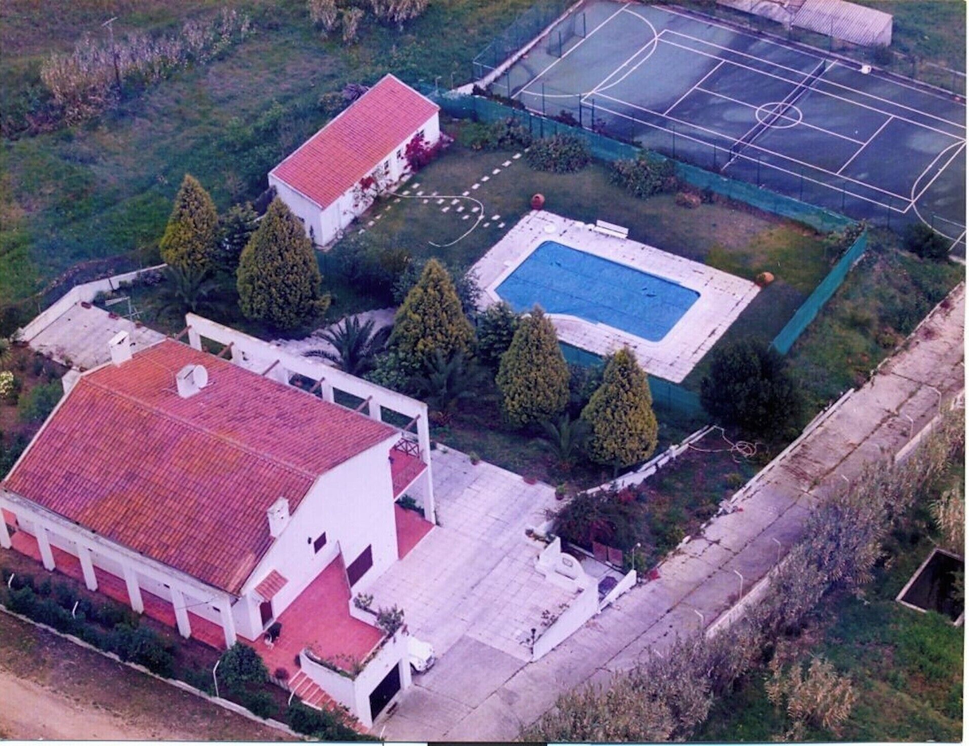 Vista aérea de una propiedad residencial con piscina, cancha de tenis y una casa grande de techo rojo.