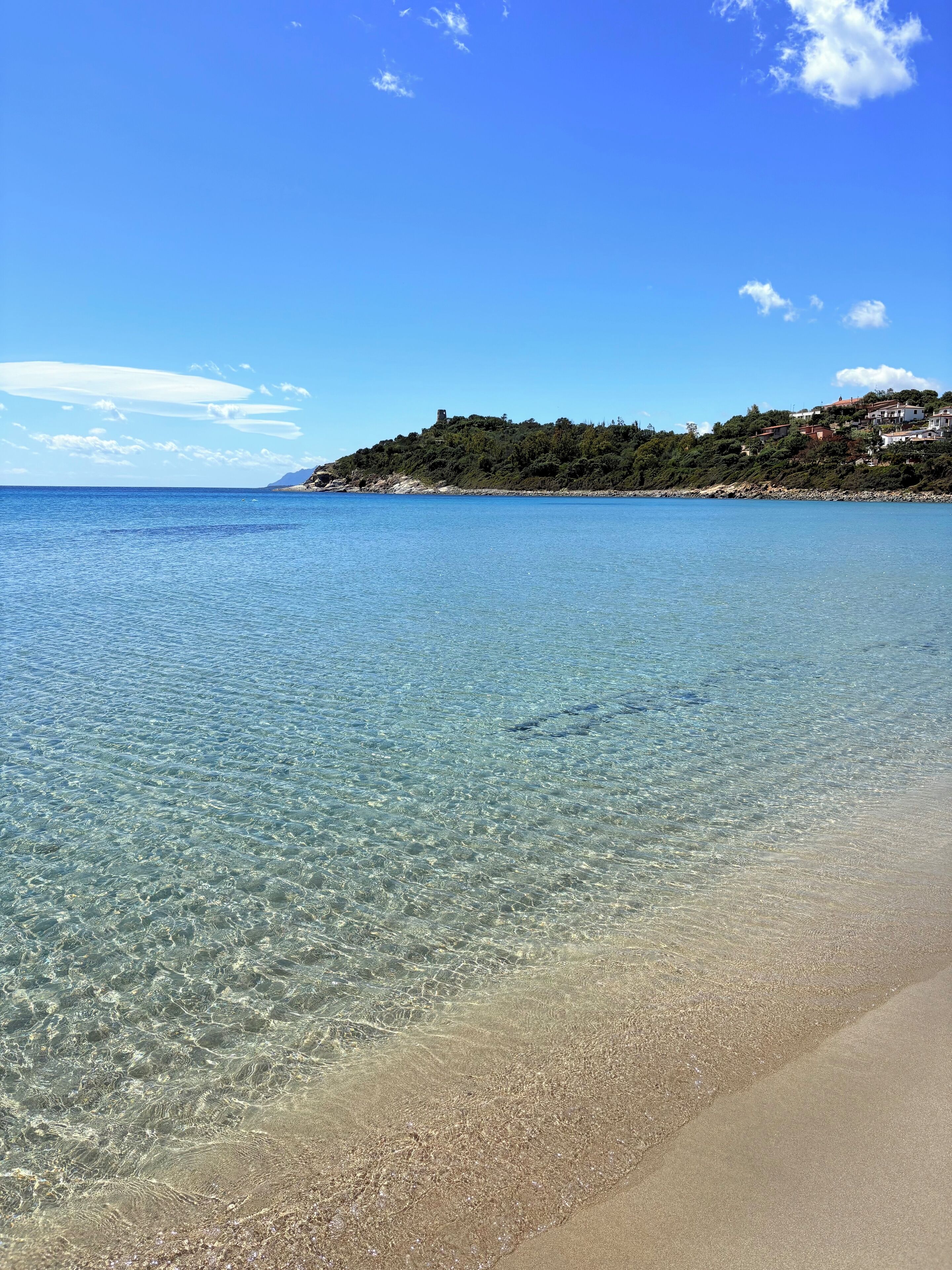 En vacker strand med grunt vatten och ett avlägset kullområde med byggnader.